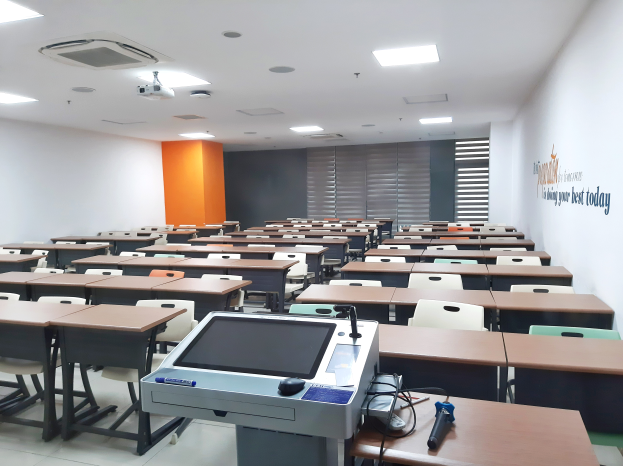 Ein Klassenzimmer mit Tischen und Stühlen in Reihen, einen deckengelagerten Projektor und verschiedene Gegenstände auf den Tischen, mit Text an der rechten Wand, der darauf hinweist, dass es sich um eine Privatschule in Singapur handelt.