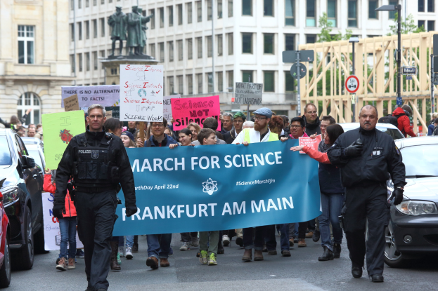 Eine Gruppe von Menschen marschiert auf einer Straße und hält ein Banner mit der Aufschrift "March for Science Frankfurt am Main" hoch, mit Autos, Gebäuden, Statuen, Laternenmasten, Schildern und Bäumen im Hintergrund.