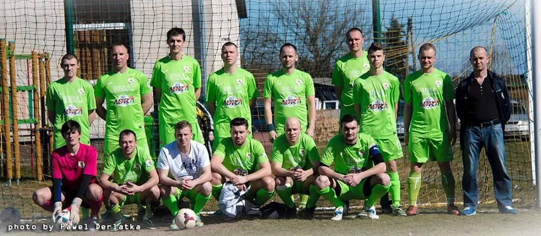 Gruppe von Männern in grünen T-Shirts auf einem Fußballfeld in der Nähe eines Torpfostens mit einem Ball auf dem Boden und verschiedenen Hintergrundelementen wie Bäume, Gebäude und Fahrzeuge.