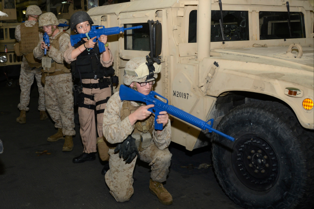 Eine Gruppe von Marines in Helmen mit blauen Gewehren steht vor einem Militärfahrzeug, mit anderen Fahrzeugen und Lichtern im Hintergrund und Text am unteren Rand des Bildes.