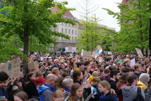 Große Menschenmenge protestiert vor einem Gebäude, hält Schilder, mit Bäumen, Fahrzeugen und einem Lautsprecher in Berlin.