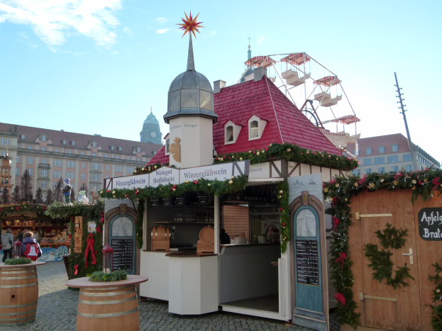 Kleines rotes Dachgebäude mit einem Riesenrad davor, umgeben von festlichen Oktoberfest-Dekorationen, Menschen und Gegenständen, vor einem Hintergrund aus Gebäuden, Bäumen und einem bewölkten Himmel.