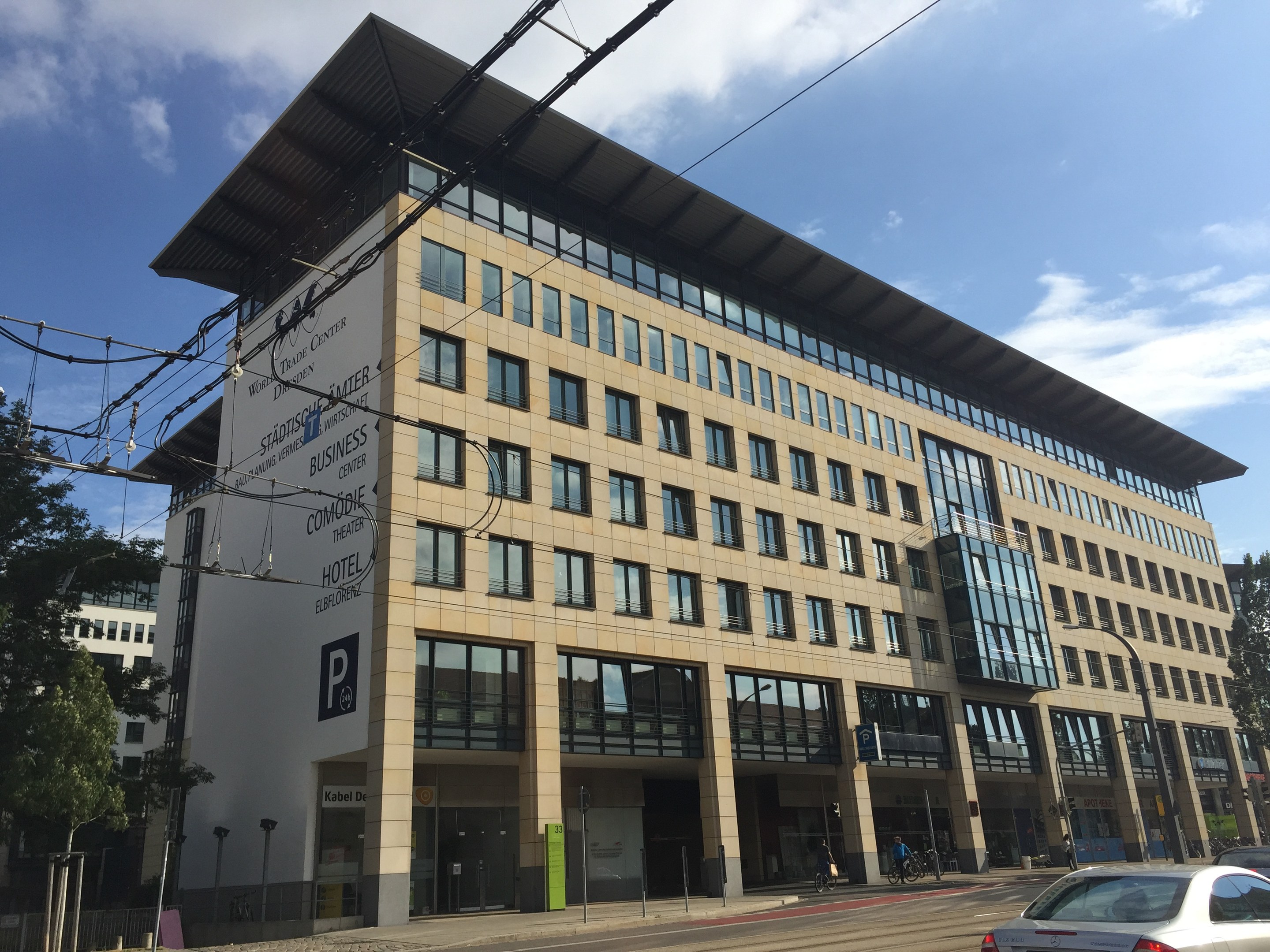 Großes modernes Gebäude mit vielen Fenstern, beschriftet mit "Stadthalle Center für Geschäft", umgeben von Bäumen und städtischer Infrastruktur, mit Menschen und Fahrzeugen im Vordergrund unter einem bewölkten Himmel.