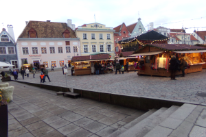 Ein belebter Weihnachtsmarkt in Tallinn, Estland mit Menschen um geschmückte St├Ąnde, festlicher Beleuchtung, bew├Âlktem Himmel, Geb├Ąuden im Hintergrund, Treppen führend nach oben und Topfpflanzen.