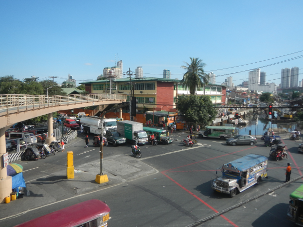 Eine belebte Stadtstraße mit Autos, Bussen und Motorrädern, eine Brücke mit Geländern und Säulen, Laternenmasten, Strommasten mit Drähten, Gebäude mit Fenstern, Bäume und ein bewölkter Himmel.