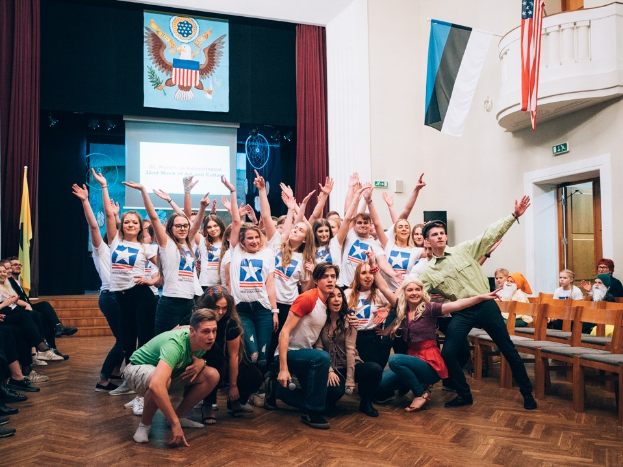 Gruppe von Menschen, die auf einem nationalen Jugendkongress-Event vor einem sitzenden Publikum posen, mit einem Bildschirm, Flaggen und anderen veranstaltungsbezogenen Elementen im Hintergrund.