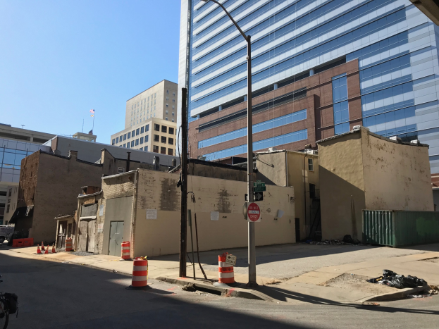 Stadtstraße mit einem geparkten Fahrrad, Gebäuden, Straßenmöblierung, Fahrzeugen, Mülltonnen und einem demolierten Gebäude im Hintergrund bei klarem blauem Himmel.