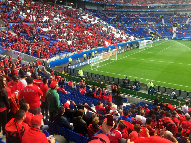 Ein volles Stadion mit Zuschauern, einige sitzen, andere stehen, die ein Fußballspiel beobachten, mit sichtbaren Torpfosten, Bannern und Deckenleuchten unter einer Dachkonstruktion.