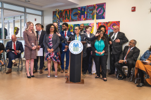 Gruppe von Menschen vor einem Rednerpult mit Mikrofon und einer Tafel mit der Aufschrift "Sancho Stadtverordneten joins Stadtverordnetenversammlung", mit sitzenden und stehenden Zuschauern, Flaggen, Feuerlöscher und Deckenbeleuchtung im Hintergrund.