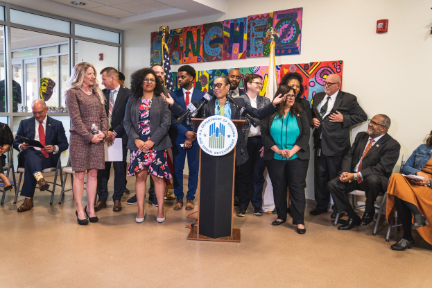 Gruppe von Menschen vor einem Rednerpult mit Mikrofon und einer Tafel mit der Aufschrift "Sancho Stadtverordneten joins Stadtverordnetenversammlung", mit sitzenden und stehenden Zuschauern, Flaggen, Feuerlöscher und Deckenbeleuchtung im Hintergrund.