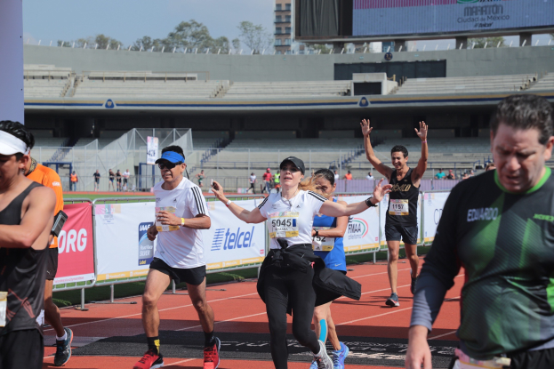 Gruppe von Menschen, die auf einer Laufbahn in einem Stadion laufen, mit Bannern, einem Metallzaun, einer Anzeigetafel, einem Gebäude, Bäumen und einem bewölkten Himmel; einige Läufer tragen Turnschuhe und Hüte.
