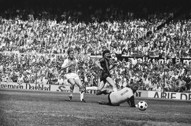 Schwarzes und weißes Foto von Männern, die auf einem Feld Fussball spielen, mit Zuschauern im Hintergrund und einer "Bundesliga 1978-79 Ajax 1-1 Hannover 96"-Fahne.