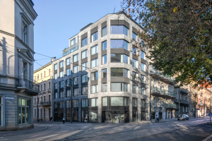 Großes Bürogebäude mit vielen Fenstern, umgeben von Straßeninfrastruktur, Fahrzeugen, Fußgängern, Bäumen und einem bewölkten Himmel, als neue Firmenzentrale identifiziert.