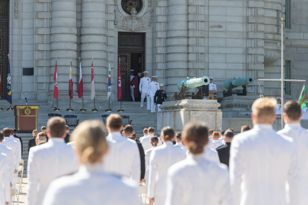 Eine Gruppe von Menschen in weißen Uniformen und Mützen steht vor einem Gebäude mit Säulen und einer Tür, mit Flaggen, einem Podium mit Mikrofon, Treppen und Kanonen im Hintergrund, was auf eine Marineakademie-Abschlusszeremonie hindeutet.