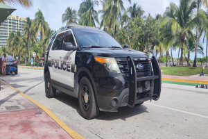 Ein Polizeiwagen am Straßenrand mit Menschen, Fahrrädern, Gras, Bäumen, Gebäuden und einem klaren blauen Himmel im Hintergrund; das Vorderrad des Wagens ist sichtbar.