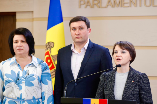 Zwei Frauen und ein Mann stehen an einem Podium vor einer ukrainischen Flagge, wobei der Mann einen schwarzen Blazer, ein weißes Hemd und schwarze Hosen trägt und die Frau rechts ein aschfarbenes Kleid trägt, während sie die Ukraine-Krise vor einer cremefarbenen Wand mit Text und einer braunen Tür links diskutieren.