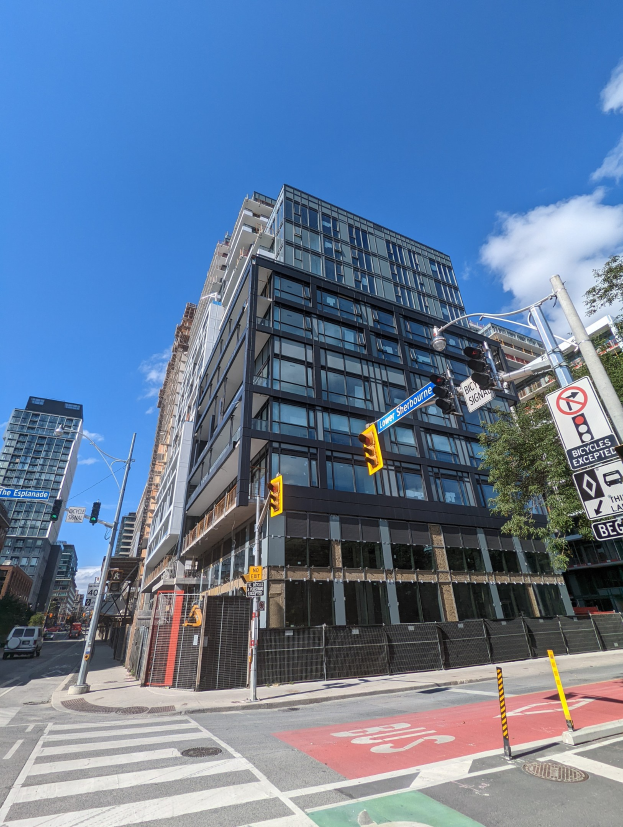 Eine belebte Stadtstraße mit einem hohen Apartment-/Reihenhaus-Gebäude im Hintergrund, Fahrzeuge auf der Straße, Verkehrsampeln, Schilder, Bäume, ein Zaun und ein bewölkter Himmel.