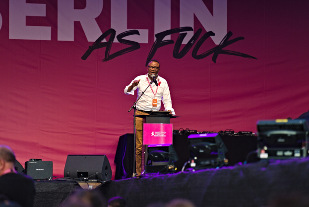 Ein Mann in einem weißen Hemd und braunen Hosen steht auf einer Bühne und hält ein Mikrofon in der Hand, während er eine Menge anspricht. Im Hintergrund ist ein pinker Banner mit der Aufschrift "Berlin as Fuck" zu sehen.