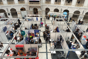 Große Gruppe von Teilnehmern auf einer internationalen Fachmesse mit Ständen, Bannern, Tischen, Stühlen und Ballons in einem Raum mit architektonischen Bögen, Säulen und einem Bildschirm im Hintergrund.