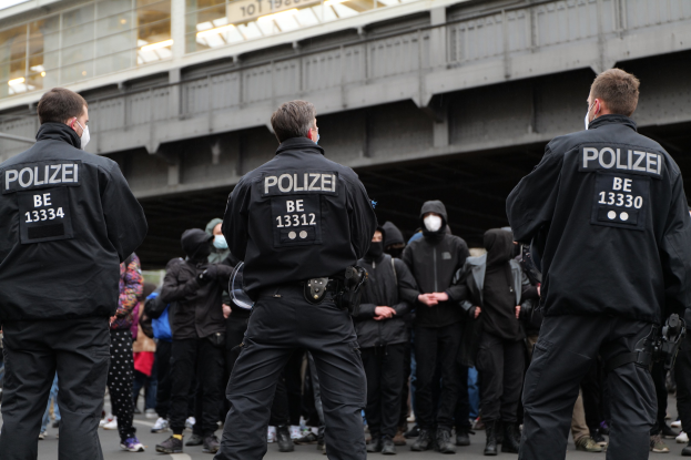 Polizisten in schwarzen Uniformen und Masken stehen vor einer Menge während einer Demonstration, mit einer Brücke und einem Gebäude im Hintergrund.