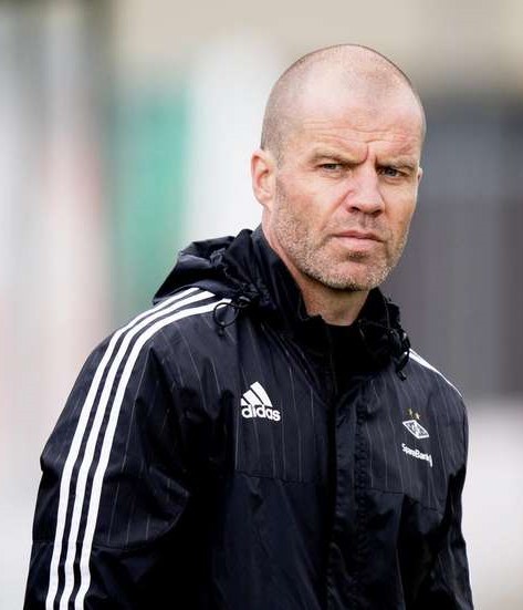 Ein kahlköpfiger Mann in einer schwarzen Jacke steht auf einem Fußballfeld, trägt eine marineblaue Jacke mit weißen Streifen und einem weißen Kragen und hat die Hände verschränkt und einen ernsten Gesichtsausdruck.