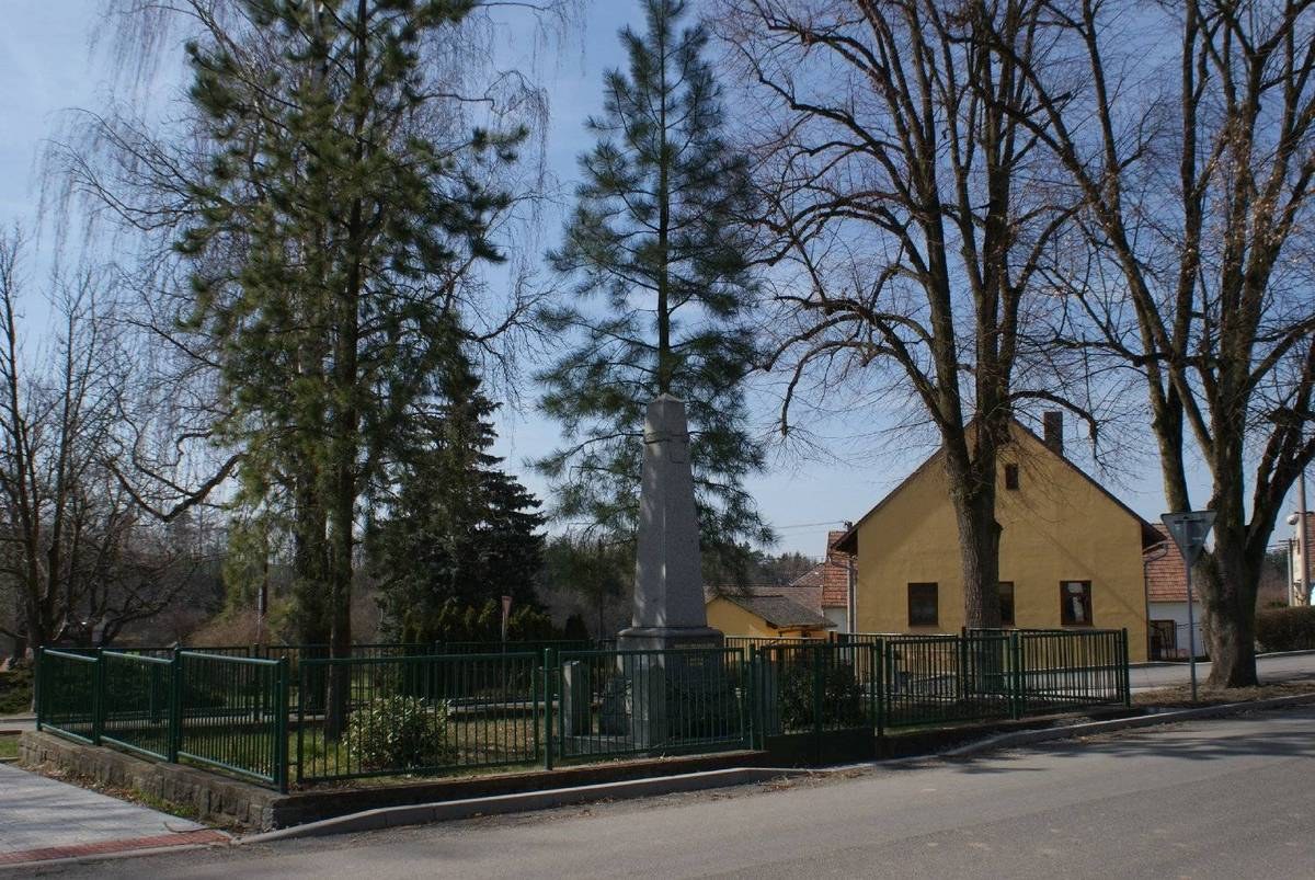 Ein Holocaust-Mahnmal steht in der Mitte einer Straße, umgeben von einem Metallzaun, umgeben von Grün, Häusern und einem bewölkten Himmel.
