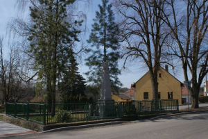 Ein Holocaust-Mahnmal steht in der Mitte einer Straße, umgeben von einem Metallzaun, umgeben von Grün, Häusern und einem bewölkten Himmel.
