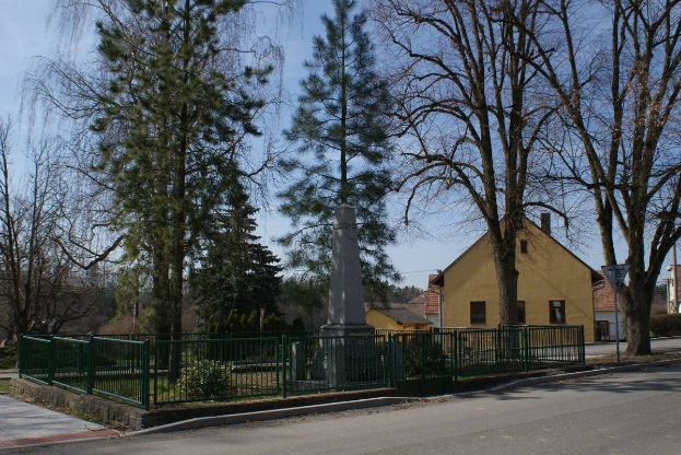 Ein Holocaust-Mahnmal steht in der Mitte einer Straße, umgeben von einem Metallzaun, umgeben von Grün, Häusern und einem bewölkten Himmel.
