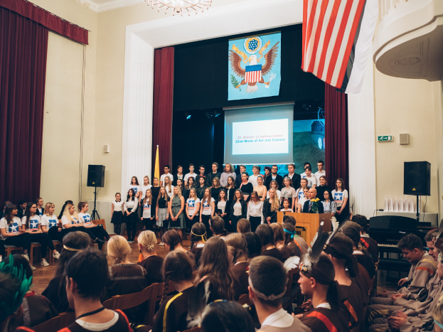 Eine Gruppe von Menschen steht vor einem sitzenden Publikum an einem Podium mit Mikrofon, flankiert von Fahnen und Lautsprechern, während eines 2. Wochen Kunst- und Kultur-Events.