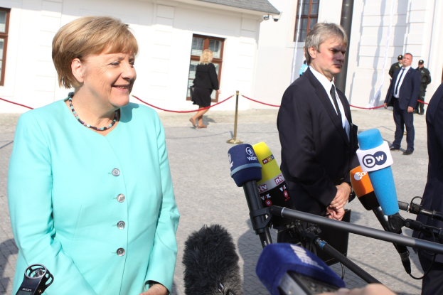 Bundeskanzlerin Angela Merkel spricht mit Reportern vor einem Gebäude mit einer Gruppe von Menschen, einige halten Mikrofonstative, mit einem Seil, das an einen Pfahl gebunden ist, unter einem bewölkten Himmel.