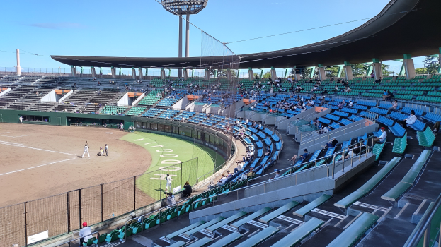 Baseball-Spiel im Gange mit Zuschauern auf den Rängen und auf dem Feld, umgeben von einem Zaun, Bäumen und einem klaren blauen Himmel im Hintergrund.