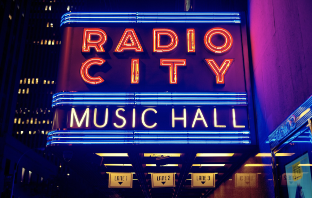 Außenansicht des Radio City Music Hall in New York City bei Nacht, mit heller Beleuchtung und hohen umliegenden Gebäuden, mit einer 'Radio City'-Leuchtschrift und zusätzlichen Schildern auf der rechten Seite.