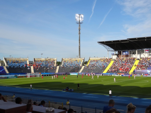 Fußballspiel in einem großen Stadion mit Spielern auf dem Feld und Zuschauern auf den Tribünen unter Flutlicht, mit dem Himmel im Hintergrund.