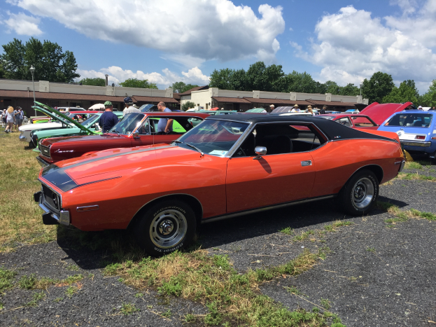 Eine Gruppe von Oldtimern auf einem Rasenparkplatz geparkt, mit Menschen in Mützen in der Nähe, Gebäuden, Bäumen und einem bewölkten Himmel im Hintergrund.