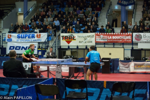 Indoor-Tischtennis-Match mit zwei Spielern und mehreren Zuschauern, die um den Tisch herum stehen und sitzen, sowie leeren Stühlen und Werbetafeln im Hintergrund.