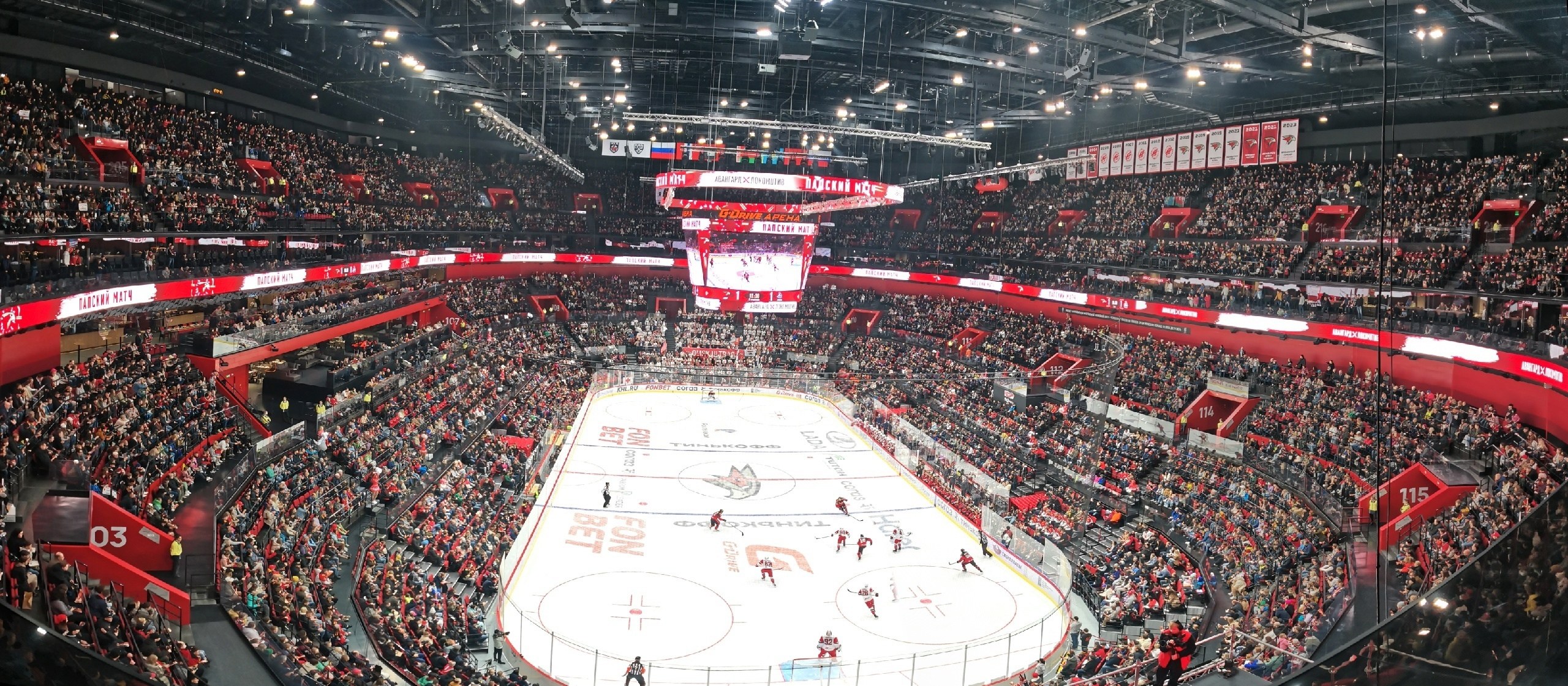 Eine große Indoor-Hockey-Halle mit Zuschauern auf den Rängen, heller Deckenbeleuchtung und einem zentralen Scoreboard-Bildschirm.