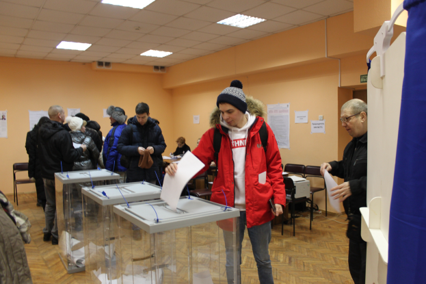 Eine Gruppe von Menschen steht um eine Wahlurne in einer Wahlkabine, einige halten Papiere, mit einem blauen Vorhang und einem weißen Objekt auf der rechten Seite, Stühlen, Tischen, Papieren an der Wand und Deckenlampen im Hintergrund.