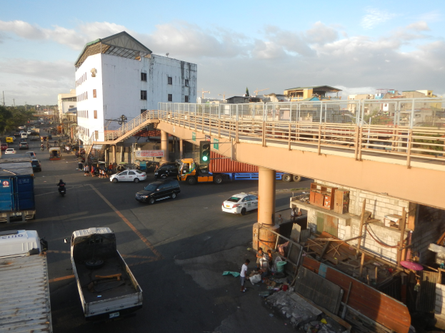 Eine belebte Stadtstraße mit Autos, Lastwagen, Fußgängern, einer Brücke mit Geländern und Säulen, Ampeln, Gebäuden mit Fenstern, Bäumen und einem bewölkten Himmel.