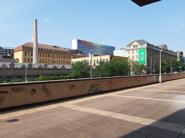 Stadtstraßenansicht mit Graffiti-bedeckter Wand, Bäumen, Straßenlaternen, einem Schild, Fahrzeugen, Gebäuden mit Fenstern, einem Schornstein und einem bewölkten Himmel.