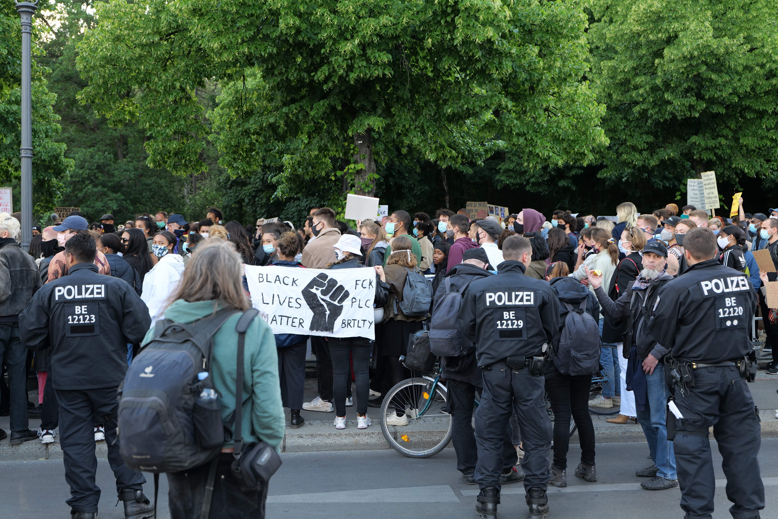 Eine große Gruppe von Menschen nimmt an einer Black Lives Matter Demonstration in Berlin teil, einige halten Schilder und tragen Mützen und Taschen, während im Vordergrund ein Fahrrad und im Hintergrund Bäume und ein Pfahl zu sehen sind.