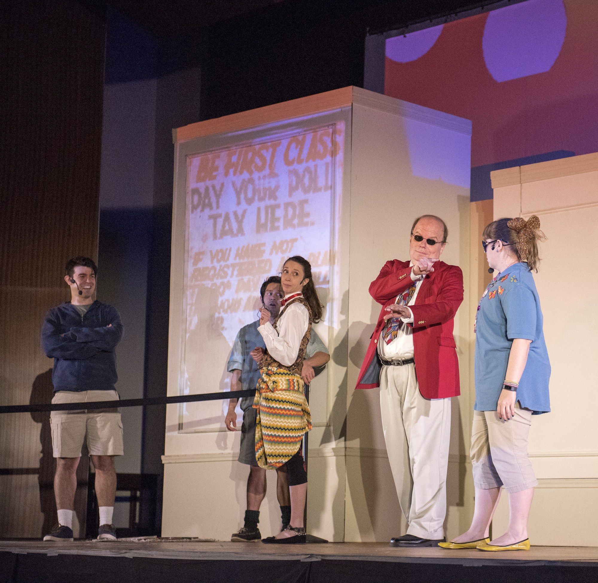 Eine Gruppe von Menschen auf einer Bühne mit einem Mann, der ein Mikrofon hält, im Zentrum, ein Display hinter ihnen zeigt "The First Class Day Your Poll Tax Here", ein Zaun, eine Wand und Deckenleuchten.