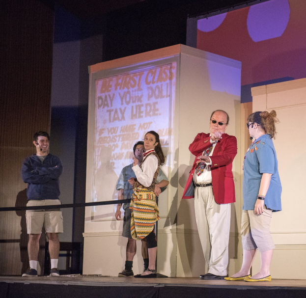 Eine Gruppe von Menschen auf einer Bühne mit einem Mann, der ein Mikrofon hält, im Zentrum, ein Display hinter ihnen zeigt "The First Class Day Your Poll Tax Here", ein Zaun, eine Wand und Deckenleuchten.