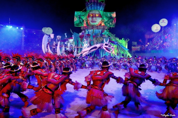 Eine Gruppe von Menschen in bunten Kostümen tanzt auf einer Bühne vor einem sitzenden Publikum, mit Lichtern, Polen, Luftballons und einem Bildschirm, der eine Person in den Hintergrund zeigt, unter einem sichtbaren Himmel, bei der Rio-Karneval 2014.