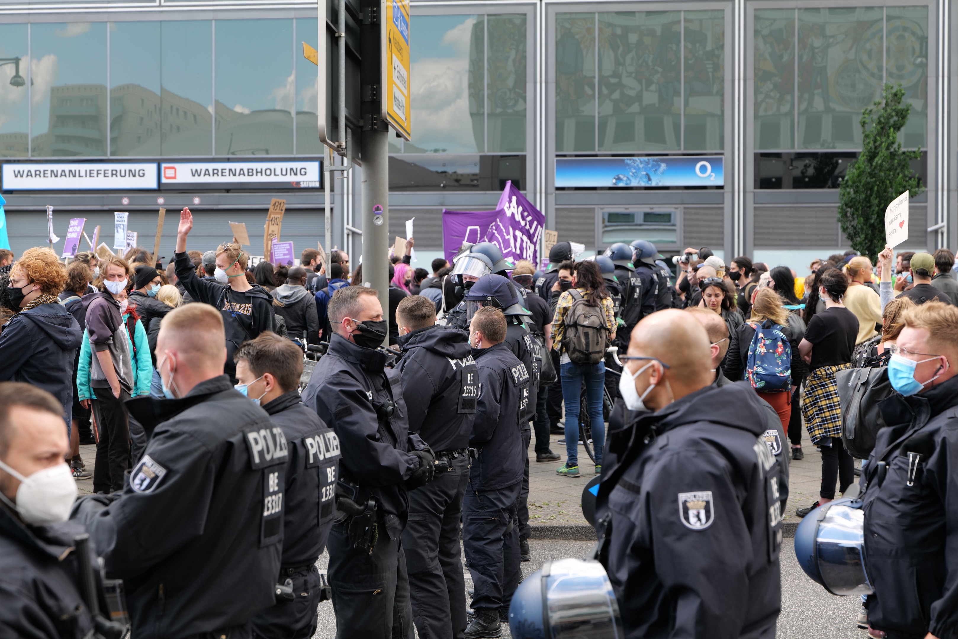 Eine große Gruppe von Menschen protestiert vor einem Gebäude, einige halten Schilder und tragen Helme, mit einem Schildständer und einem Baum in der Nähe.