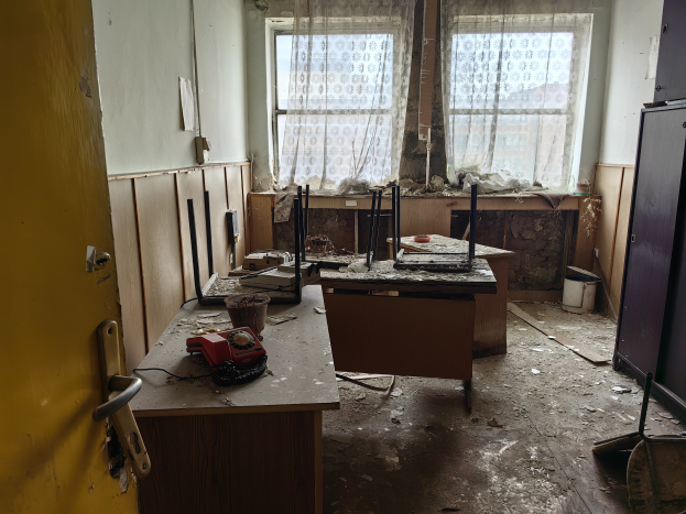 Leerer Raum mit einer Tür auf der linken Seite, einem Schrank auf der rechten Seite und einem Fenster mit Vorhängen im Hintergrund; ein zentraler Tisch hält verstreute Gegenstände, die auf ein verlassenes Büro hindeuten.