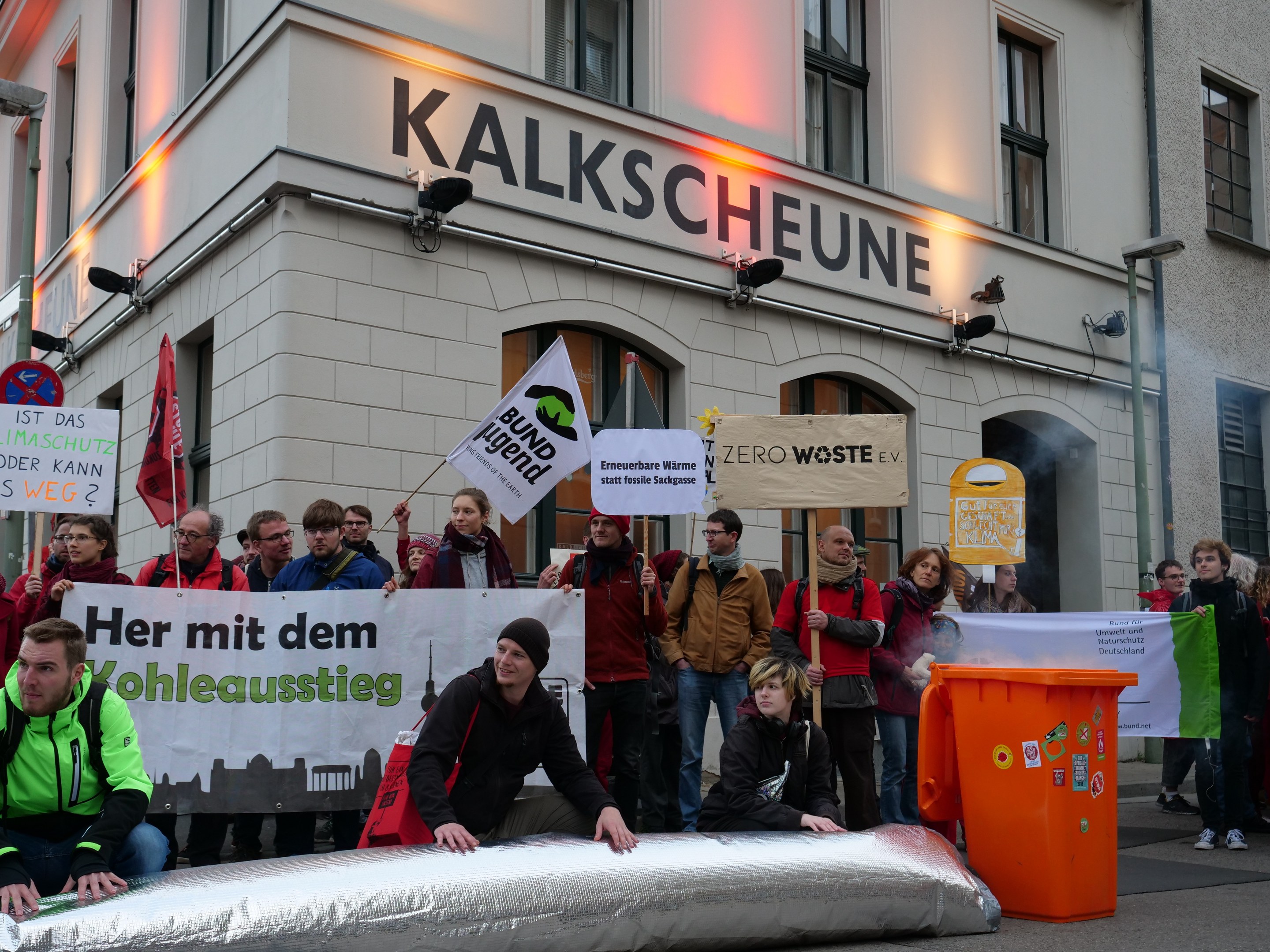 Gruppe von Menschen mit Protestschildern und Plakaten vor einem Gebäude, mit zwei Personen im Vordergrund und einem Müllcontainer auf der rechten Seite.
