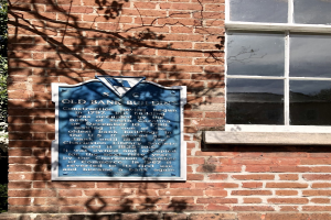 Ein altes Bankgebäude mit einer blauen Plakette an der Seite, ein Fenster auf der rechten Seite und einen Baum auf der linken Seite.