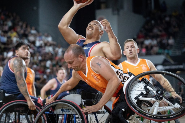 Eine Gruppe von Männern in Rollstühlen beim Basketballspielen, einer hält den Ball, mit Zuschauern im Hintergrund.