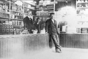 Drei Männer stehen an einem Tresen in einem Geschäft mit verschiedenen Gegenständen, während ein Regal mit Büchern und anderen Gegenständen im Hintergrund zu sehen ist, während offenbar ein Raubüberfall stattfindet.