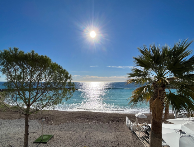 Ein sonniger Strand mit Palmen, Schirmen und üppiger Vegetation, vor einem strahlend blauen Himmel mit der Sonne im Hintergrund.